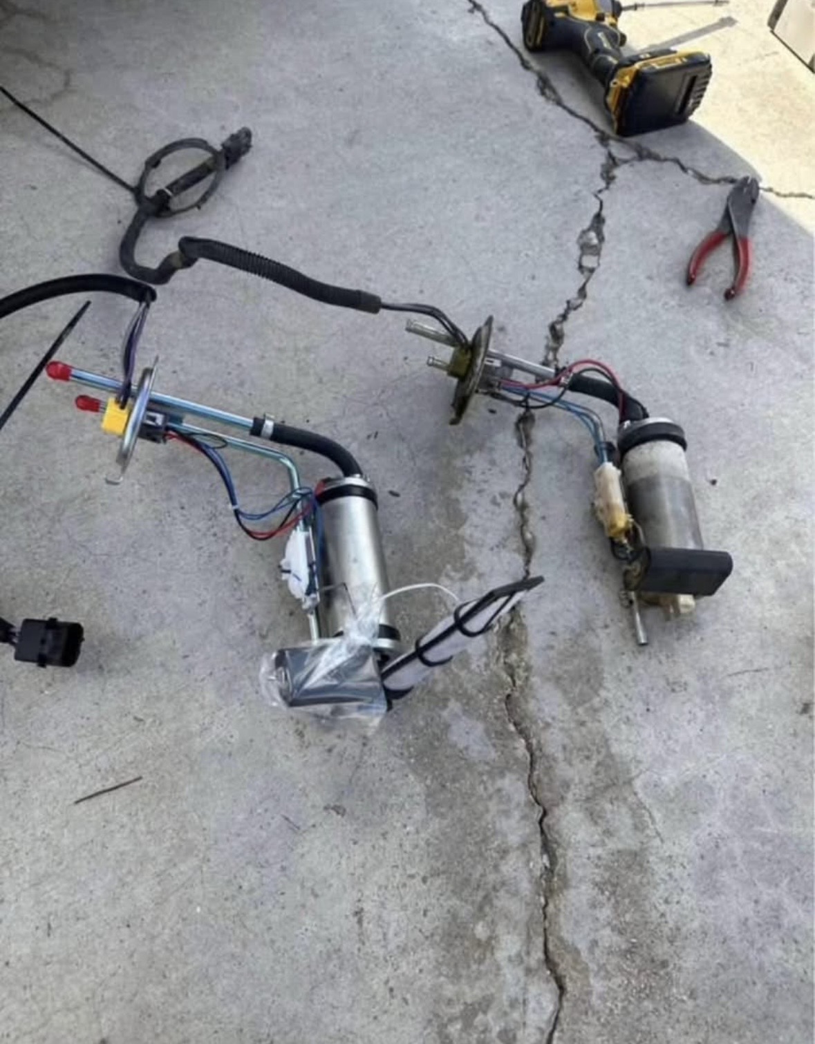 Two fuel pump assemblies laid out on concrete beside a cordless drill and pliers