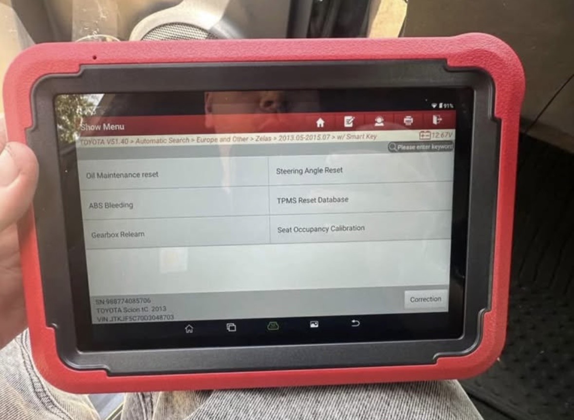 Hand holding a red diagnostic scan tablet showing a calibration and reset menu inside a car
